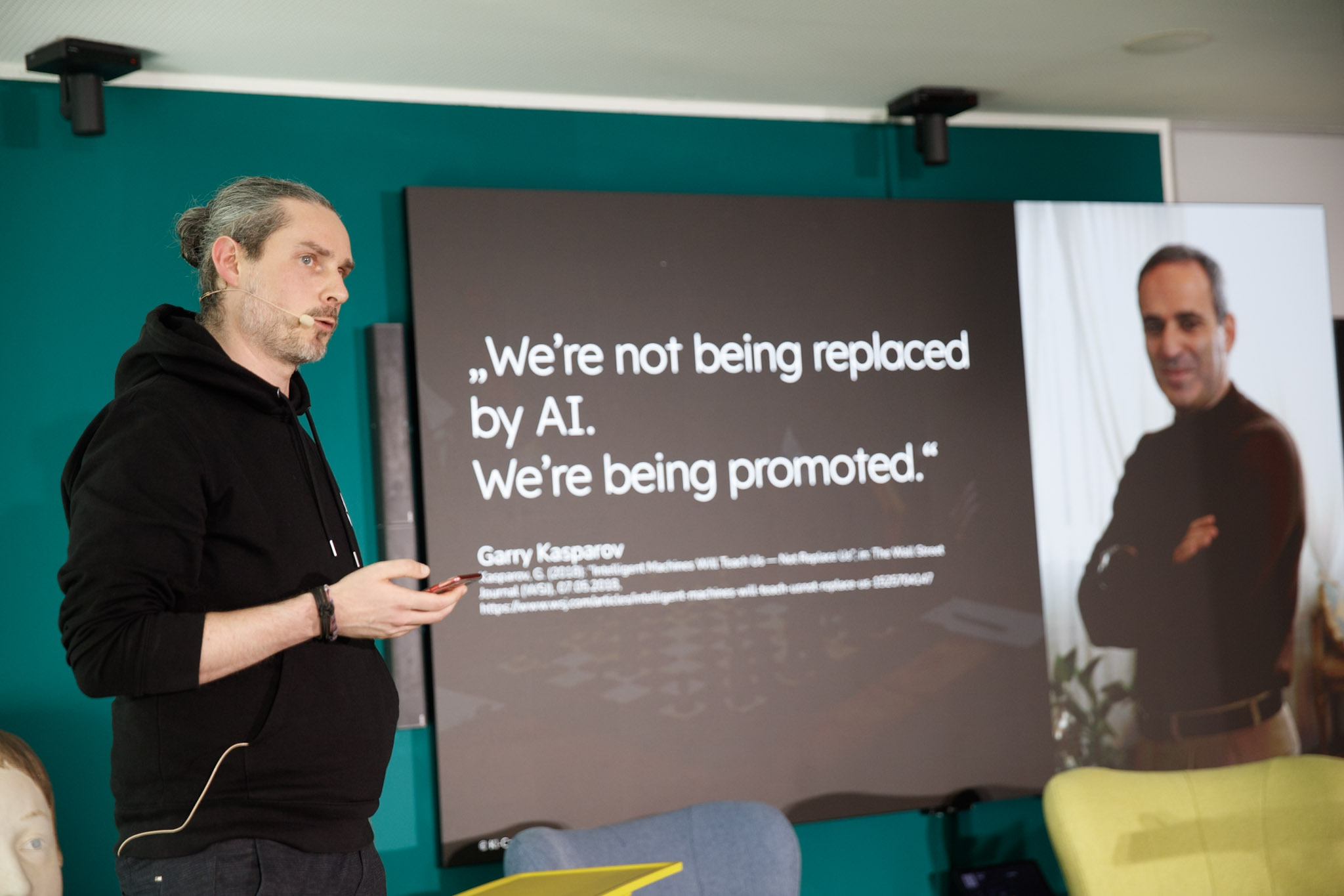 ki-weiterbildung-schulung-agentur Christian Kleemann steht auf einer Bühne und gibt eine KI-Schulung und Weiterbildung. Er zeigt auf eine Leinwand mit einem Zitat zu KI von Garry Kasparov auf dem steht: We're not being replaced by AI. We're being promoted.